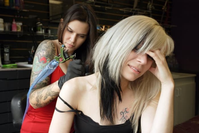 Nainen tatuointitaiteilija, joka soveltaa käsityötään 30 -vuotiaiden naisten selkään ja käsivarteen. (Kiinteistöjulkaisu tatuointikuville liitteenä)