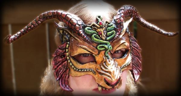 CHIMERA MASK En maske baseret på det mytiske monster, Chimera, i rigt guld, orange og rødt. Traditionelt havde kimæren en løvekrop, en løve og gedehoveder og en slangehale. Kunstneren gav masken hovedet på en hornet løve med en slange, der slingrede ned i trynen og gedemblemet på panden. Ged og slange har skinnende røde øjne, og en rød dråbe er placeret i slangens pande. Masken er helt håndskulptureret og malet, som har et kunstnerstempel på bagsiden med datoen den blev oprettet. Masken er let og hornene er noget fleksible for at reducere risikoen for skader. Med horn er mærket 14